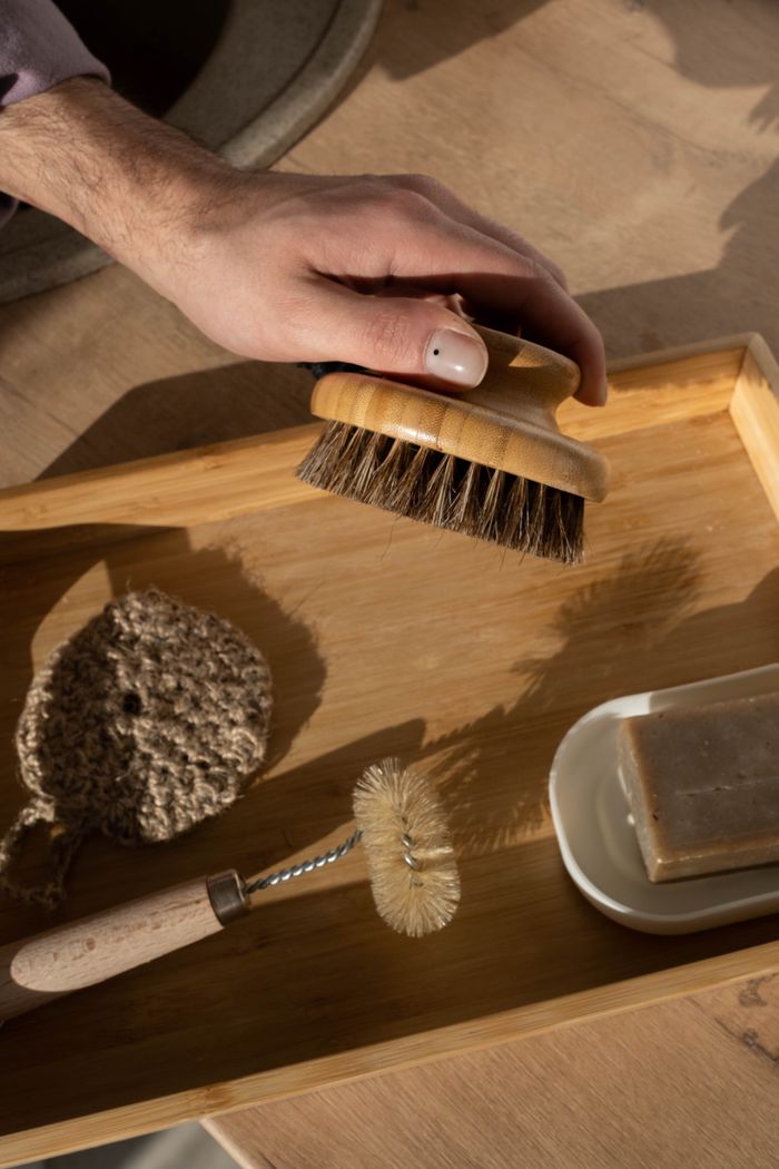 hero-02 Close-up of hand holding a wooden brush on a bamboo tray with cleaning tools.